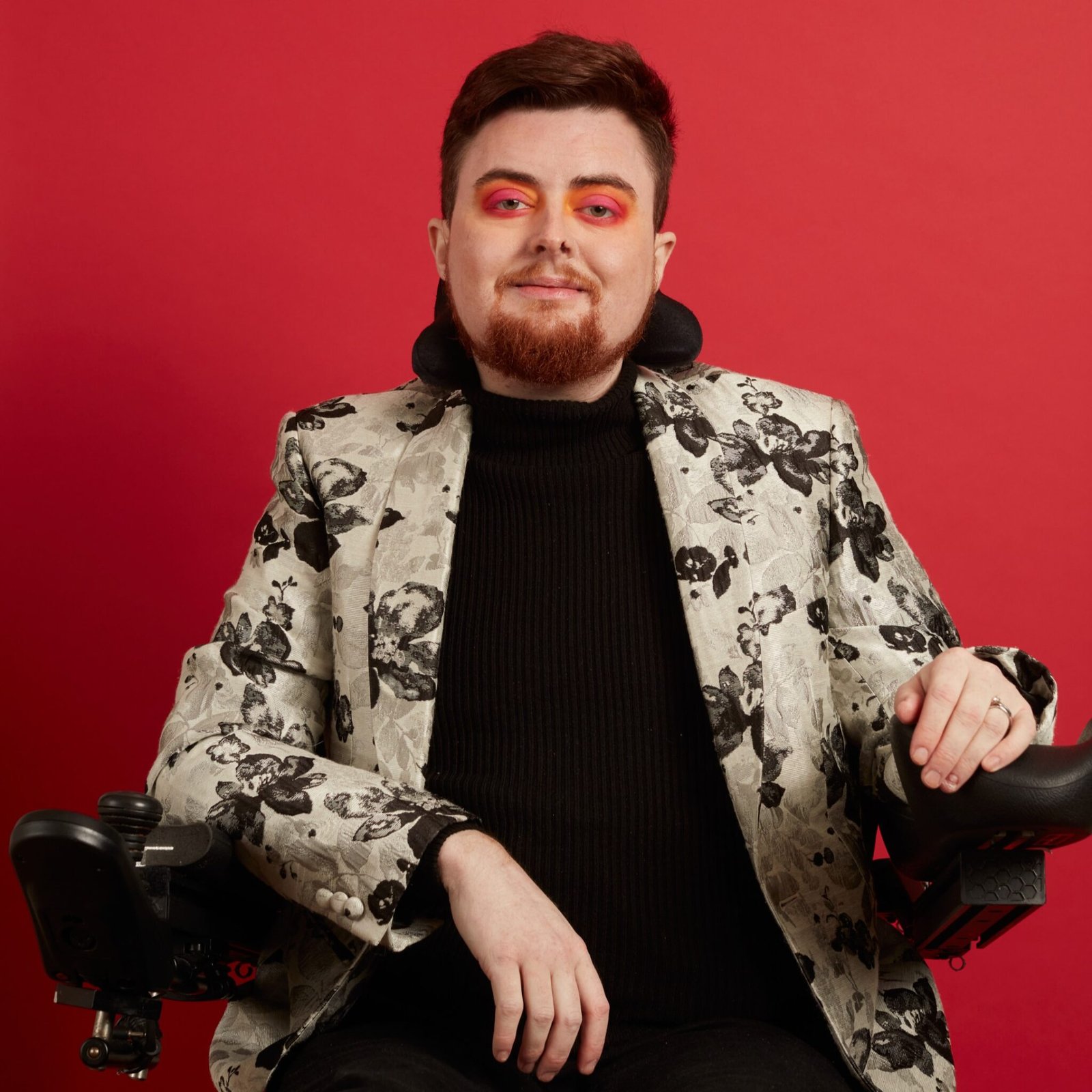 Jamie Hale by Holly Falconer. Jamie is a white person in an electric wheelchair with dark red hair and beard, wearing a black top and silver blazer. The background is red and their eyeshadow is orange.
